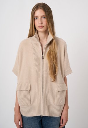 Junge Frau mit langen, glatten Haaren trägt einen beigen, kurzärmeligen, gestrickten Zip-up-Pullover mit zwei Fronttaschen und blauen Jeans.