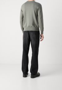 Henkilö, joka wearing vaaleanharmaan neuletakin, mustat farkut ja mustat kengät, seisoo selkä kameraan päin yksinkertaista valkoista taustaa vasten.