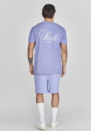 Mann trägt lilafarbenes SikSilk T-Shirt und passende Shorts, weiße Socken und weiße Turnschuhe, steht mit dem Rücken zum Betrachter vor neutralem Hintergrund.