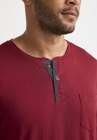 T-shirt in cotone rosso con scollatura arrotondata, patta dei bottoni accentuata in grigio e una piccola tasca anteriore. Finitura testurizzata con tessuto a coste.