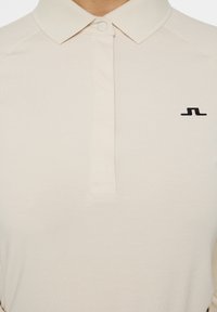 Beige Poloshirt aus glattem Stoff mit Knopfverschluss, geripptem Kragen und schwarzem Logo auf der linken Seite.