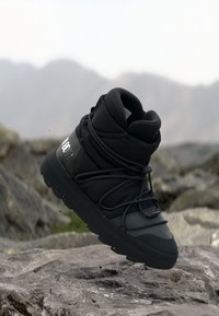 Botă de iarnă izolată, neagră, cu talpă groasă și șireturi din sfoară, suspendată deasupra unui teren stâncos, cu un fundal montan neclar.