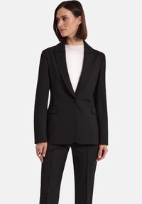 Kobieta ubrana w czarną dopasowaną marynarkę z jednym guzikiem, zestawioną z pasującymi czarnymi spodniami i białą bluzką z okrągłym dekoltem, na białym tle.