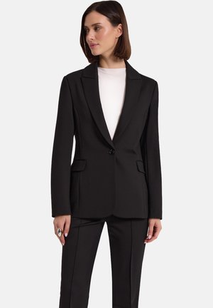 Kobieta ubrana w czarną dopasowaną marynarkę z jednym guzikiem, zestawioną z pasującymi czarnymi spodniami i białą bluzką z okrągłym dekoltem, na białym tle.