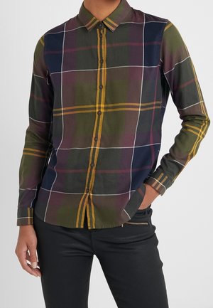 Person wearing a fitted button-up shirt with large olive, navy, and mustard plaid pattern, paired with black pants, one hand in pocket.