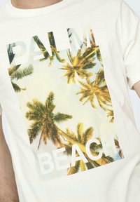 Camiseta de color crema claro con un gráfico de una palmera, que presenta las palabras "PALM" y "BEACH" en letras blancas de bloque a través del diseño.