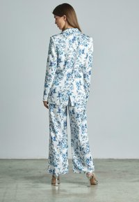 Costume à motifs floraux en blanc et bleu, composé d'un blazer ajusté et de pantalons évasés. Tissu lisse, coupe droite et talons argentés.