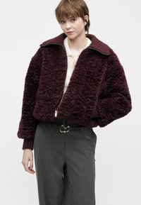 Junge Person, die eine burgunderfarbene, strukturiertete Zip-Jacke über einem cremefarbenen Oberteil, graue Hosen mit einem schwarzen Gürtel mit einer goldenen Schnalle trägt.