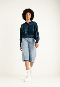 Pulover s pomladnimi prugami v mornarsko modri in zeleni barvi, s ovratnikom, v kombinaciji z svetlo modrimi jeans culottes in belimi športnimi copati, na enobarvni podlagi.