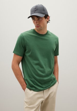 Green cotton t-shirt with a crew neck, paired with light beige trousers. A grey cap with a letter "P" accentuates the outfit.