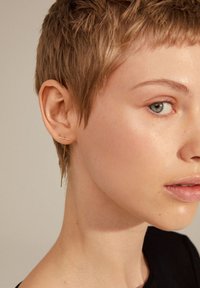 Orecchini color oro presentano una sottile barra orizzontale e un delicato contorno verticale, riflettendo la luce su uno sfondo neutro.