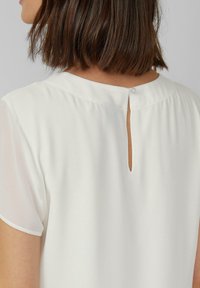 Frau mit kurzen braunen Haaren, die eine weiße Bluse mit einem kleinen Rückenausschnitt trägt, der mit einem einzelnen Knopf verschlossen ist.