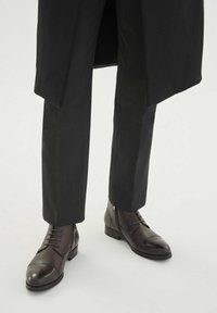 Rjavi usnjeni škornji z vezalkami, zaobljenim nosom, nizkim petom in subtilnimi detajli šivov, kombinirani z temnimi krojenimi hlačami.