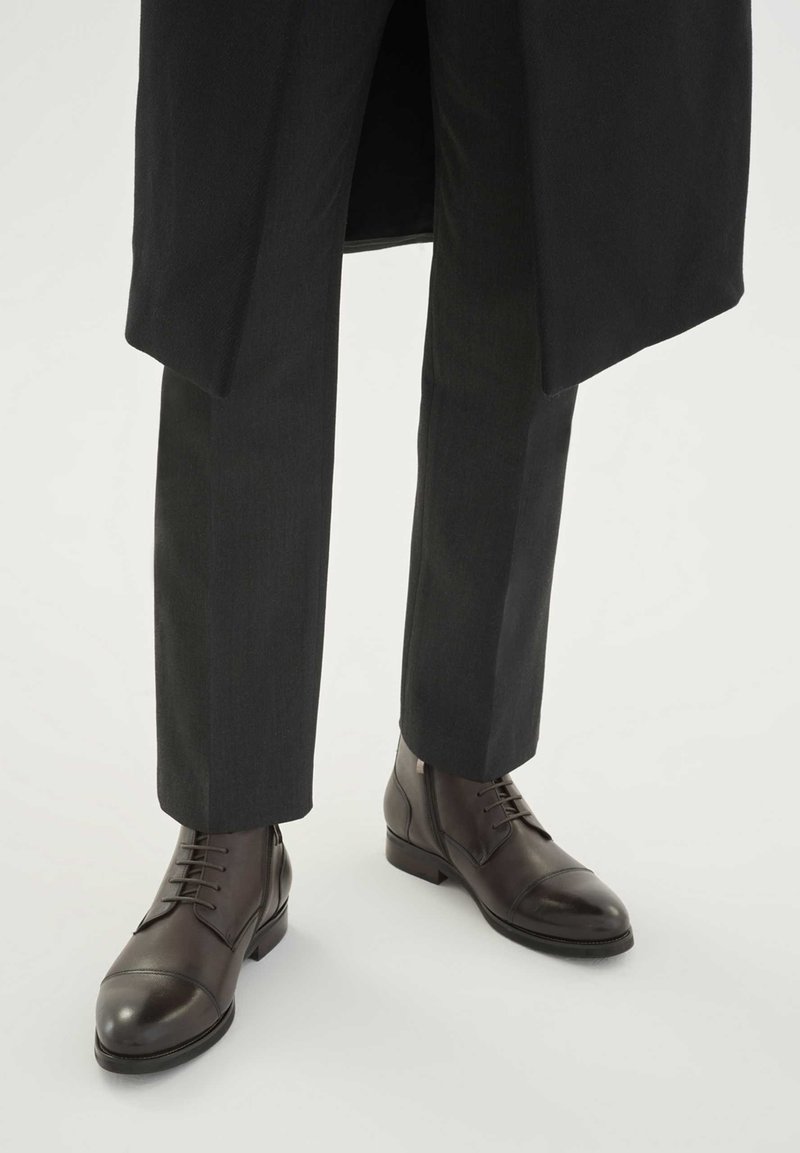 Rjavi usnjeni škornji z vezalkami, zaobljenim nosom, nizkim petom in subtilnimi detajli šivov, kombinirani z temnimi krojenimi hlačami.