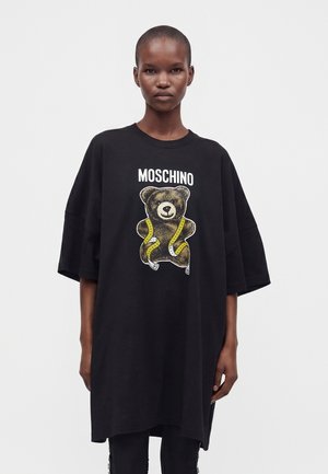 Lühikeste juustega noor inimene kannab üle suuruse musta Moschino T-särki, millel on kollase mõõdulindiga ümber mässitud kaisukaru.