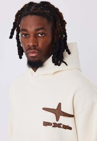 Hoodie color crema realizzato in tessuto morbido, caratterizzato da una stella cucita marrone e un design del logo sul lato sinistro del petto. Vestibilità comoda con cappuccio regolabile.