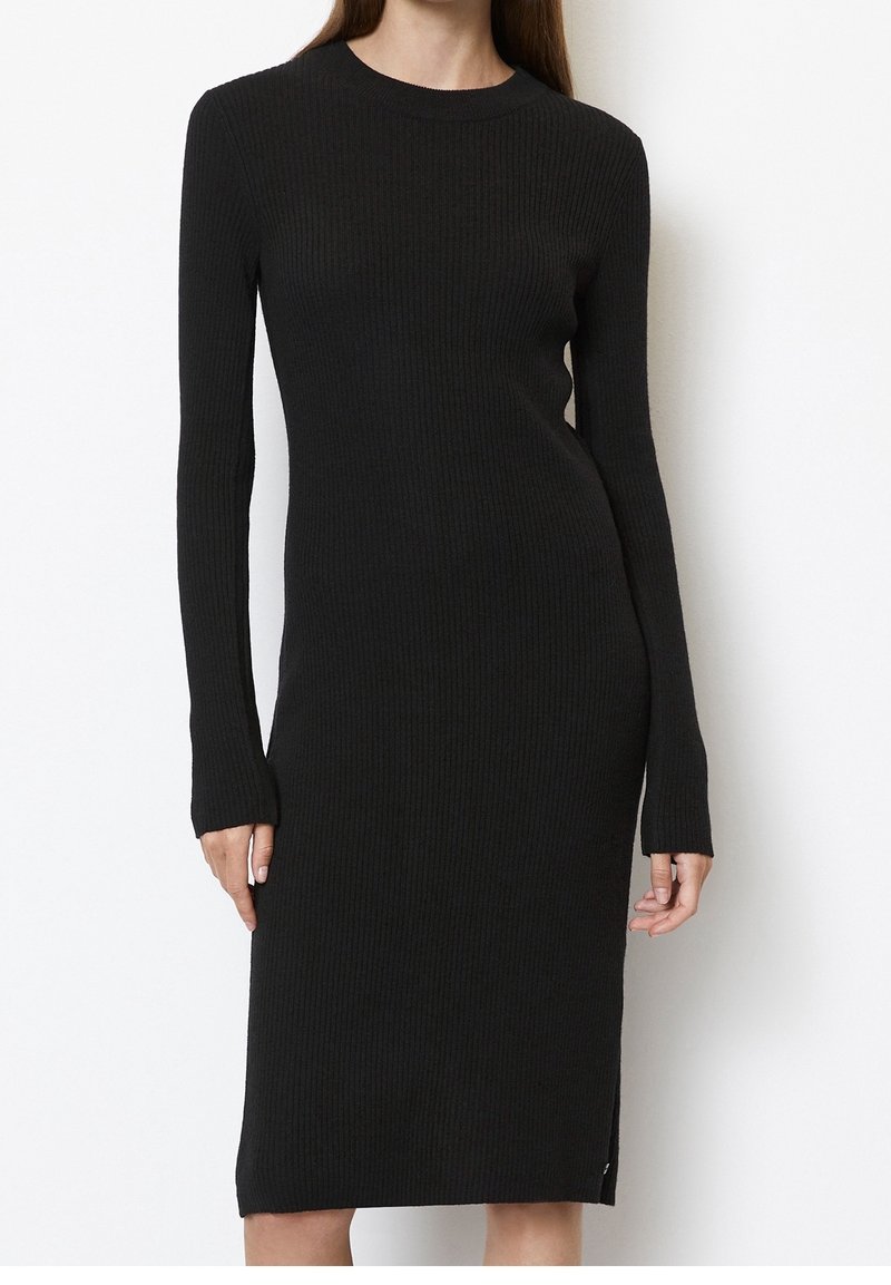 Femme portant une robe noire en maille côtelée, ajustée, à manches longues et longueur genou, sur fond blanc.
