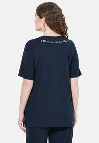 Námořnická modrá tričko s krátkými rukávy, s zakulaceným spodním lemem. Na zadní straně je bílý text a grafika ryb s nápisem "ponoř se s proudem."