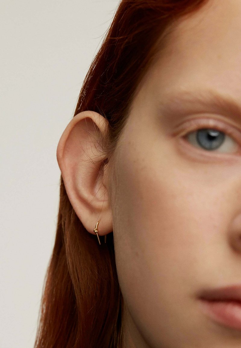 Pendiente de aro dorado con un diseño delicado, que presenta un pequeño acento sobre un fondo claro, usado en la oreja izquierda de una persona.