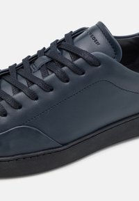 Tmavě modré kožené tenisky s texturovaným povrchem, plochou černou gumovou podrážkou a tkanicemi. Obsahují decentní logo na jazyku.