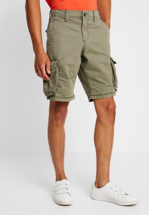 Mand iført olivengrønne cargoshorts, hvide sneakers og en orange skjorte, stående med hænderne i lommerne mod en ensfarvet baggrund.