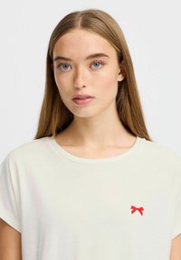 Cremefarvet t-shirt med korte ærmer har en lille rød sløjfe broderi på venstre brystområde. Glat stof og afslappet pasform.