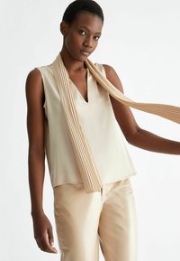 Top beige senza maniche con scollo a V, abbinato a pantaloni in pelle beige a vita alta. Una sciarpa plissettata si appende elegantemente attorno al collo.