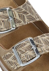 Sandeal beige con doble hebilla, con una correa con un diseño de hojas. La plantilla parece acolchada y se presenta sobre un fondo neutro.