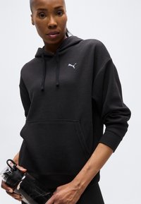 Γυναίκα που φοράει μαύρο φούτερ Puma κρατώντας ένα μαύρο παγούρι σε απλή ανοιχτή φωτεινή επιφάνεια.