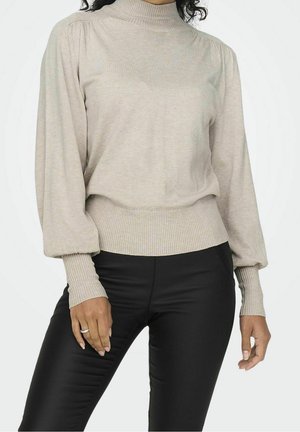 Beiger Pullover mit hohem Kragen, langen Puffärmeln und gerippten Bündchen, getragen über schwarzen, figurbetonten Hosen. Weicher Stoff, minimalistischem Design.