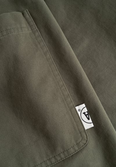 Gros plan sur un tissu vert avec une poche cousue et une étiquette blanche portant un logo circulaire noir partiel et du texte.