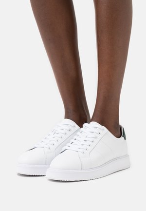 Dark-skinned person wearing white low-top lace-up sneakers with a green heel tab, standing on a white surface.