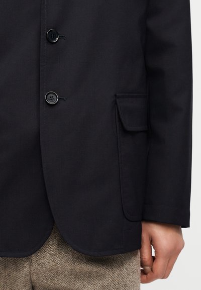 Κοντινό πλάνο ενός ατόμου που φοράει σκούρο μπλε σακάκι με μαύρα κουμπιά και τσέπη με καπάκι, σε συνδυασμό με καφέ υφασμένα παντελόνια.
