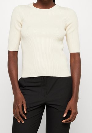 Kvinde iført en tætsiddende, ribstrikket cremefarvet kortærmet sweater kombineret med højtaljede sorte bukser, stående foran en ensfarvet baggrund.