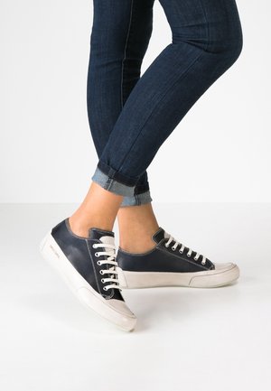Persoon die donkerblauwe opgerolde jeans en marineblauw-witte veterschoenen draagt, staand met gekruiste benen op een witte vloer.