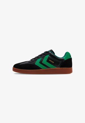 Schwarze und grüne Sneaker aus Wildleder und synthetischem Leder, mit Chevron-Mustern und einer braunen Gummisohle. Goldene Schrift an der Seite.
