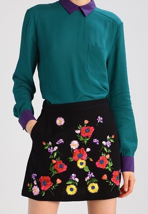 Mujer que lleva una camisa de manga larga color verde azulado con cuello y puños morados, combinada con una falda negra bordada con coloridos patrones florales.