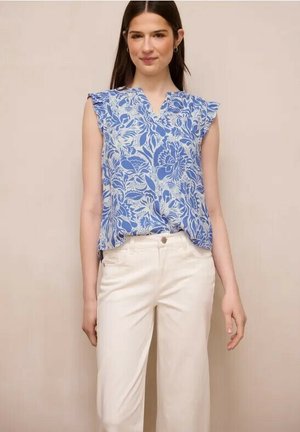 Kvinde iført ærmeløs blå blomstret bluse med flæseærmer og hvide bukser med høj talje, stående mod beige baggrund.