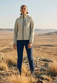Femme debout sur un terrain rocheux avec de l'herbe sèche, portant une veste beige et un pantalon bleu marine, faisant face à l'avant avec un paysage désertique en arrière-plan.
