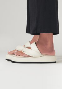 Sandali platform bianchi con design a strap annodato, presentano una texture morbida e una suola piatta in gomma nera. Indossati con pantaloni neri.