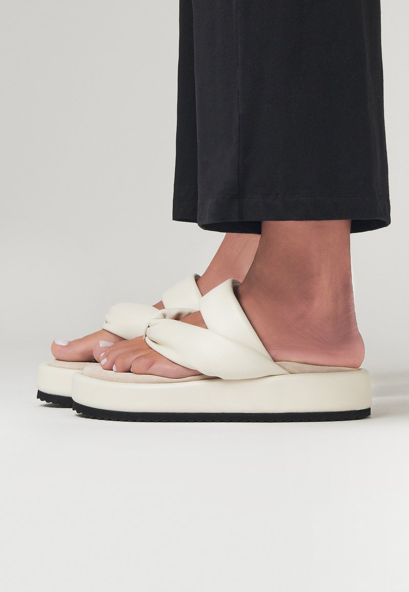 Sandali platform bianchi con design a strap annodato, presentano una texture morbida e una suola piatta in gomma nera. Indossati con pantaloni neri.