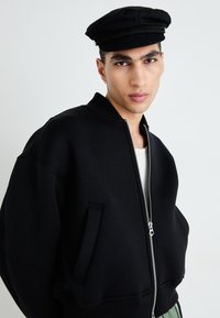 Veste bomber noire avec fermeture éclair à l'avant, poignets côtelés et poches latérales. Assortie d'une casquette noire avec un accent tressé.