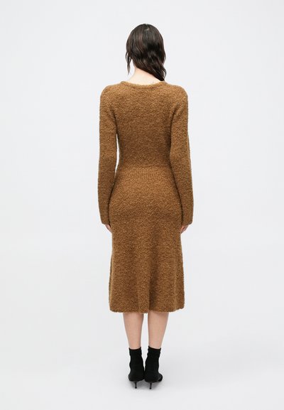 Bruine jurk met lange mouwen, gemaakt van een gestructureerd, pluizig materiaal, aangesloten aan de taille en uitlopend tot onder de knie. Draagt zwarte enkellaarsjes.