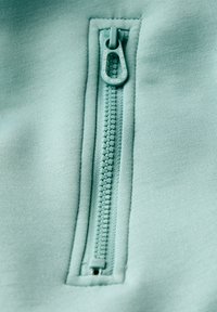 Detailní záběr na světle tyrkysový zip na látce, se stahovákem označeným "CECIL" a vzájemně propletenými plastovými zuby podél zipového pásu.