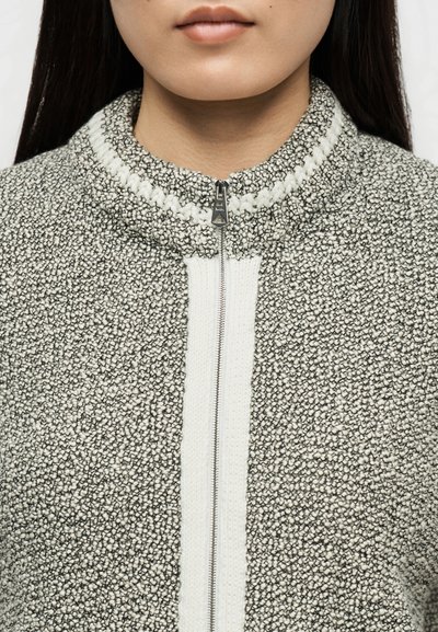 Γυναίκα που φοράει γκρι και άσπρο πλεκτό πουλόβερ με φερμουάρ και ψηλό γιακά, με εμφανή τα χείλη και το κάτω μέρος του προσώπου.