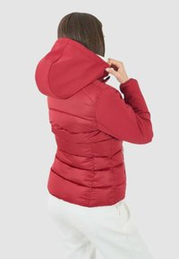 Giacca imbottita rossa con cappuccio, caratterizzata da cuciture orizzontali e una silhouette aderente. Trama morbida e tasche laterali con zip.