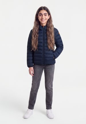 Teenagepige med langt, bølget hår iført en mørkeblå dunjakke, grå jeans og hvide sneakers, stående og smilende med den ene hånd i lommen.