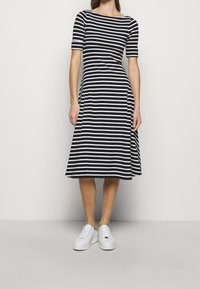 Woman wearing black and white horizontal striped knee-length dress and white sneakers on a plain light gray background.