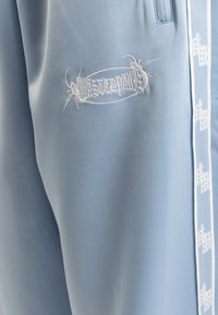Light blue joggers made of smooth fabric featuring white embroidered text and an insect motif on the front and white side stripes.