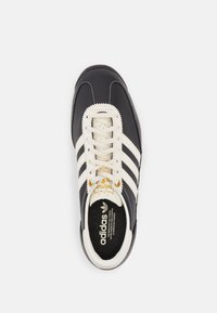 Adidas-tennis kengät tummansinisestä nahasta, kermaiset nauhat ja kolme erottuvaa kermaista raitaa. Teksturoitu musta pohja ja kultainen logoyksityiskohta.